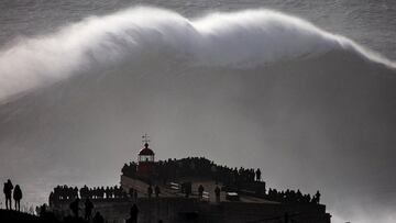 La ola gigante de Nazaré, con surfistas en el agua, rompiendo frente al Fuerte de San Miguel Arcángel (en Nazaré, Portugal), con mucho público viendo un evento de surf tow-in.