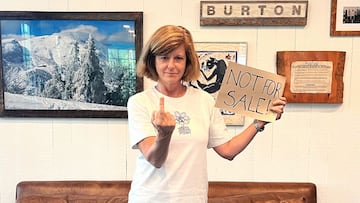 La presidenta de Burton Snowboards, Dona Carpenter, sostiene un cartel de Not for sale con una mano y con la otra realiza una peineta. Frente a un sofá y unos cuadros.