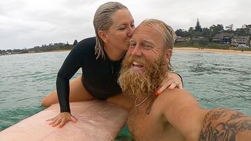 El surfista Blake Johnston con su pareja en el mar, sobre una tabla de surf, haci&eacute;ndose un selfie con la playa al fondo. 