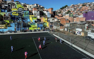 Residentes jugando al fútbol en un campo con la imagen del jugador de fútbol brasileño Gabriel Jesús en el fondo, pintado en las paredes de las casas de la favela donde vivió durante su infancia en las afueras de Sao Paulo.