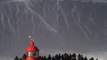 El surfista alemán Sebastian Steudtner surfeando una ola gigante en Praia do Norte, Nazaré (Portugal) el 18 de enero del 2018. Con el faro rojo del fuerte de San Miguel Arcángel en primer plano y gente mirando.