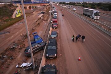 La gente camina junto a los automóviles y camiones que se encontraban entre los escombros arrastrados por las recientes inundaciones repentinas a lo largo de la carretera V-31 cerca del municipio de Massanassa el 1 de noviembre de 2024 en las afueras de Valencia, España.