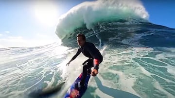 Kai Lenny grab&aacute;ndose sobre su tabla de surf con el palo selfie de la GoPro en la ola de Nazar&eacute; (Portugal). Detr&aacute;s suyo se ve el labio de la ola rompiendo justo sobre &eacute;l y un cielo despejado con el sol iluminando el agua. 