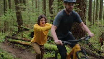 Un hombre vestido de amarillo que simula hacer el papel del motor el&eacute;ctrico de una e-MTB amarilla de Trek, la Fuel EXe, empujando a un ciclista en un bosque con muchos &aacute;rboles. 