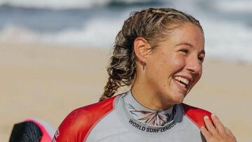 La surfista australiana de 14 a&ntilde;os Willow Hardy, con la licra de la World Surf League de color gris y rojo, sonriendo, con trenzas, en la playa, con la tabla de surf bajo el brazo. 