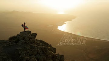 Caleta de Famara y la playa de Famara vistos desde el Risco de Famara, con un surfista con su tabla de surf en lo más alto. Lanzarote.