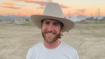 El freeskier Kyle Smaine sonriendo con un sombrero de vaquero y una camiseta blanca, en un terreno de tierra durante la puesta de sol. 