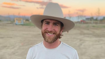 El freeskier Kyle Smaine sonriendo con un sombrero de vaquero y una camiseta blanca, en un terreno de tierra durante la puesta de sol.