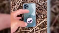 Una mano recoge un tel&eacute;fono m&oacute;vil del suelo en el campo, con ramas de fondo. 