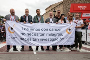 El ministro del Interior en funciones, Fernando Grande-Marlaska (2i) sujeta una pancarta que reza: 'Los niños con cáncer no necesitan milagros, necesitan investigación' antes de comenzar la etapa de montaña.