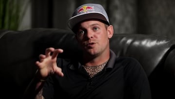 El skater Ryan Sheckler hace el gesto de la garra con la mano, con un polo negro, gorra Red Bull y sentado en un sofá negro.