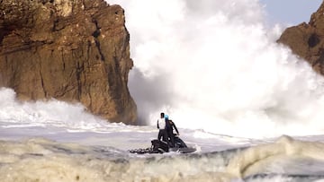 Un Jet Ski con un piloto y un fotógrafo se mete entre las rocas de los acantilados de Praia do Norte (Nazaré, Portugal) mientras rompe una ola gigante y deja una gran cantidad de espuma blanca frente a ellos, un día de noviembre del 2022.