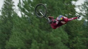 El piloto canadiense de Mountain Bike Freeride, vestido de rojo y con casco Red Bull, soltando una mano del manillar en un momento de su v&iacute;deo de X Games Real MTB, con &aacute;rboles al fondo. 