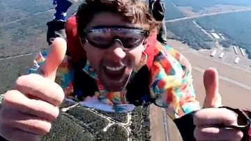 Travis Pastrana, vestido con una camisa de colores, levanta los pulgares y sonr&iacute;e a la c&aacute;mara durante un salto en paraca&iacute;das sobre una zona de bosque. 