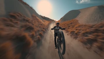 El piloto de MTB Kilian Bron, en un plano frontal, montando en MTB a una rueda por un sendero de la Capadocia (Turquía) con arena a ambos lados, arbustos y el sol al fondo.