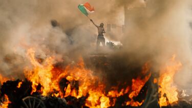 Un hombre ondea una bandera mientras el humo y las llamas se elevan desde un vehículo en llamas durante una protesta contra las redadas federales de inmigración, en el centro de Los Ángeles.