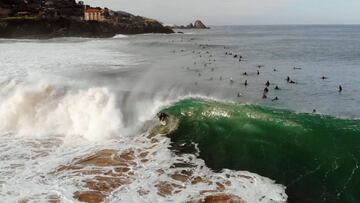 Un surfista surfeando una ola en forma de tubo de izquierdas en Mundaka (Vizcaya, País Vasco, España), con las montañas al fondo y una larga cadena de surfistas remontando hacia el pico, el 20 de diciembre del 2020.