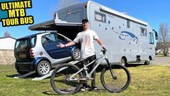 El ciclista Sam Pilgrim con su bici de MTB y su autocaravana detr&aacute;s, con un coche Smart entrando en ella. 