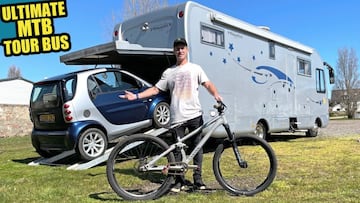 El ciclista Sam Pilgrim con su bici de MTB y su autocaravana detr&aacute;s, con un coche Smart entrando en ella. 