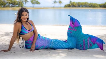 La sirena Elle Jimenez posando con su aleta frente a una playa de arena blanca con palmeras. 