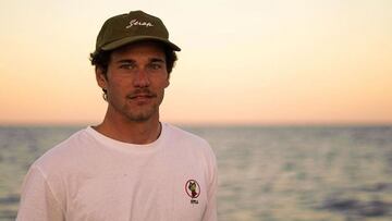 El surfista valenciano Oscar Serra, con una camiseta blanca y una gorra, mirando a cámara con el mar de fondo al atardecer.