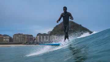Desde el agua, el surfista de origen senegalés Ousmane Sabaly 'Peter' da pasos sobre su tabla de longboard surf en una ola que rompe en la playa de la Zurriola (San Sebastián, País Vasco, España), con la playa, edificios y el Monte Urgull de fondo y un cielo parcialmente nublado.