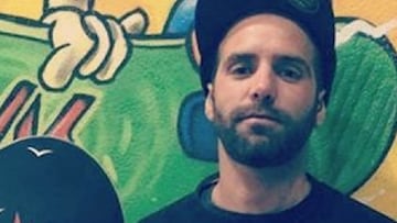 El skater Quim Monter, con gorra y con su tabla de skate, frente a una pared pintada con un graffiti en el que hay una mano agarrando una tabla de skate verde.