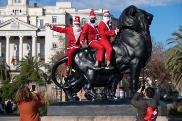 Tres participantes vestidos con trajes de Papá Noel subidos a una estatua de un león en el Puerto de Barcelona.