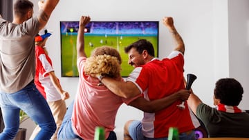 Group of young friends having fun watching football match on TV, drinking beer and cheering; football fans watching game at home celebrating after their team scoring a goal