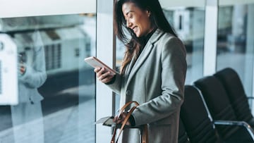 Woman standing at airport lounge looking at her mobile phone and smiling. Business traveler at airport waiting for the flight.