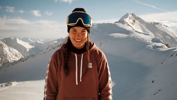 La snowboarder Maria Hidalgo, en sudadera, gorro y m&aacute;scara, mirando a c&aacute;mara y sonriendo con el glaciar de Stubai (Austria) lleno de nieve, de fondo. 