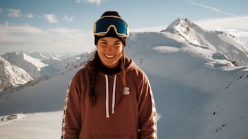 La snowboarder Maria Hidalgo, en sudadera, gorro y máscara, mirando a cámara y sonriendo con el glaciar de Stubai (Austria) lleno de nieve, de fondo.