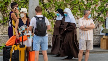 Unos turistas esperan el autob&uacute;s mientras una pareja de monjas de la Orden de Santa &Aacute;ngela de la Cruz  pasan por detr&aacute;s con mascarillas el d&iacute;a que se ha anunciado la ca&iacute;da de un 98,9% de turistas extranjeros en Andaluc&i