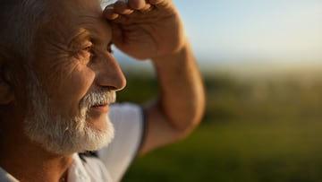 Senior bearded man using hand to protect eyes from sun, smiling outside. Close up view of male tourist squinting from bright light, while looking into distance, during mountain trip. Concept of watch.