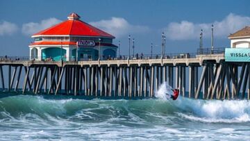 Un surfista con licra roja surfea una ola en Huntington Beach (California) con el embarcadero de fondo.