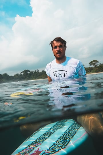 Natxo Gonzalez esperando olas en el line up con una camiseta blanca y su tabla de surf.