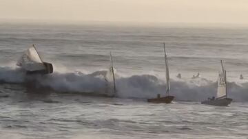 Una ola grande y brusca en marea alta golpea a 4 veleros con niños que se dirigían al puerto de Santa Cruz (California, Estados Unidos) en enero del 2021. Al fondo, un grupo de surfistas aguarda también olas para surfear.