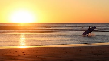 Un surfista pasea con su tabla de surf a contraluz en una puesta de sol en una playa de California (Estados Unidos).