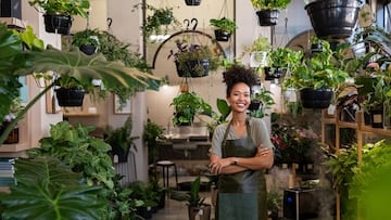 Portrait of african american woman with crossed arms wearing apron standing in botanical store. Smiling young woman in botany store standing between plants looking at camera. Happy small business owner working at flower shop standing surrounded by plants.