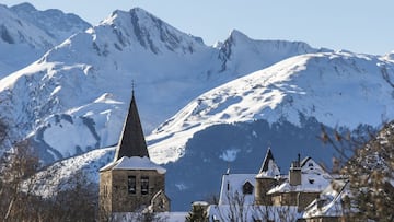 Un pueblo de la Val d&#039;Aran nevado, en primer plano, con &aacute;rboles secos. Con las monta&ntilde;as del Valle de Ar&aacute;n al fondo, tambi&eacute;n nevadas. Cielo despejado.
 