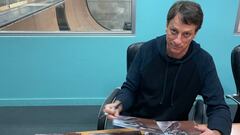 El skater estadounidense Tony Hawk firmando p&oacute;sters con su foto patinando, mirando a c&aacute;mara, con una pared azul al fondo con ventana a su Vert indoor. 