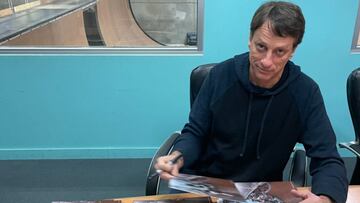 El skater estadounidense Tony Hawk firmando p&oacute;sters con su foto patinando, mirando a c&aacute;mara, con una pared azul al fondo con ventana a su Vert indoor. 