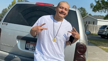 Nathan Apodaca hace el símbolo de la victoria con las dos manos, camiseta blanca y un colgante frente a su coche gris, con matrícula de Idaho.