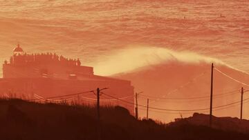 Ola grande rompiendo en Nazar&eacute; frente al faro y a mucha gente mirando, al atardecer, con colores rojos y naranjas.