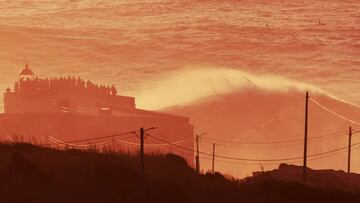 Ola grande rompiendo en Nazaré frente al faro y a mucha gente mirando, al atardecer, con colores rojos y naranjas.