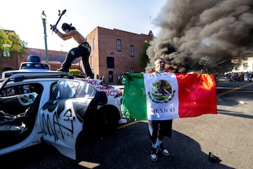 Un manifestante golpea un auto autónomo de Waymo con su patineta mientras otro se quema por detrás, durante una protesta contra las redadas federales de inmigración, en el centro de Los Ángeles.