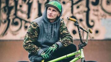 El rider de BMX Rhys Kember sobre su bici, con guantes, gorra, capucha y un graffiti al fondo.