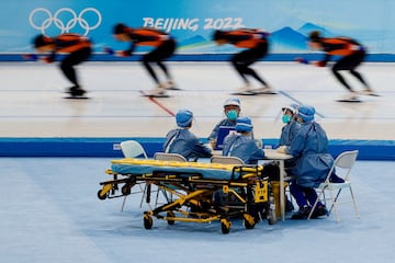 El personal médico con equipo de protección personal sentado en una mesa junto a una camilla durante una sesión de entrenamiento de patinaje de velocidad en los Juegos Olímpicos de Invierno de Beijing 2022. 
