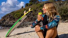 Stephanie Gilmore comiendo una manzana en la playa, con su tabla de surf al fondo.