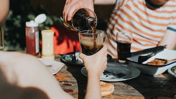 A child holds a glass out towards a man, who fills it with a fizzy drink. The scene is warm and sunny, and the drink looks refreshing. The table has an array of barbecued food around the glass.